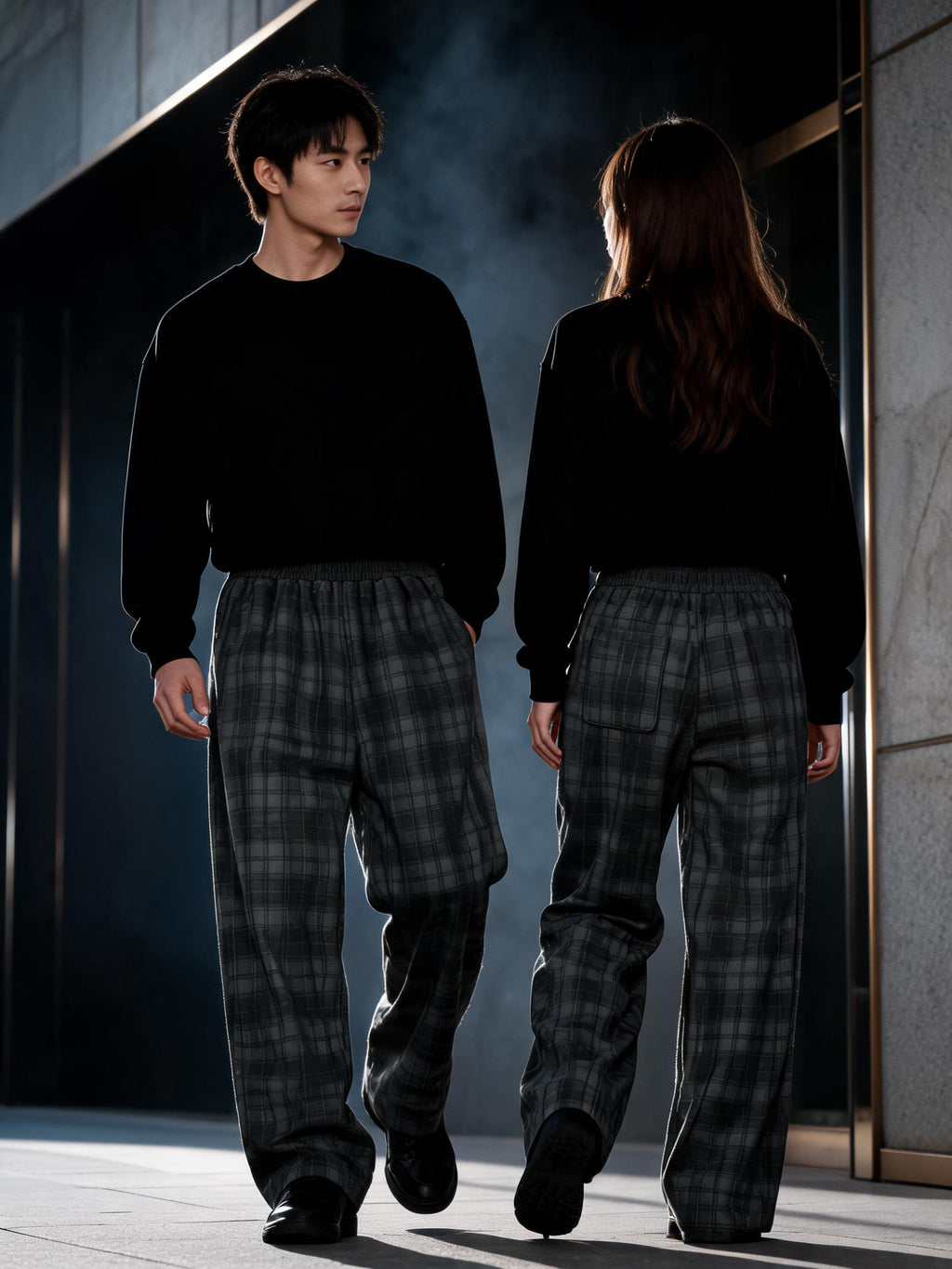 A man and a woman walking away from the camera in a dark, modern corridor. Both are wearing matching black crewneck sweatshirts and loose, wide-leg plaid pants in dark gray and black.