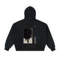 A black pullover hoodie shown from the back, featuring a large rectangular graphic print with a dripping ink design in black and beige, alongside small white text, on a plain white background.
