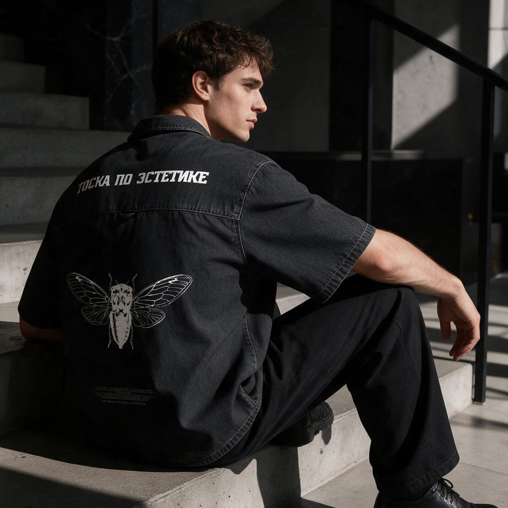 A man sits on concrete stairs in a modern building, wearing a black short-sleeve shirt with white text and a white moth graphic on the back, paired with black pants and black shoes.
