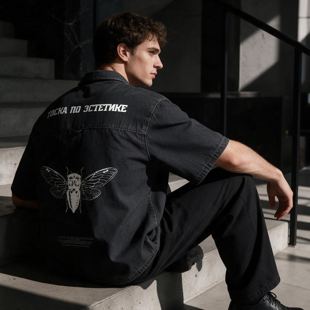 A man sits on concrete stairs in a modern building, wearing a black short-sleeve shirt with white text and a white moth graphic on the back, paired with black pants and black shoes.