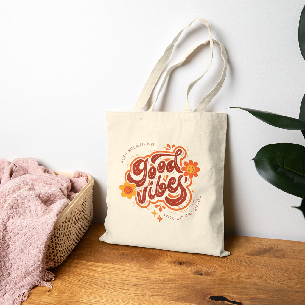 Beige tote bag with 'Good Vibes' design on a wooden surface with a white wall and plant in the background