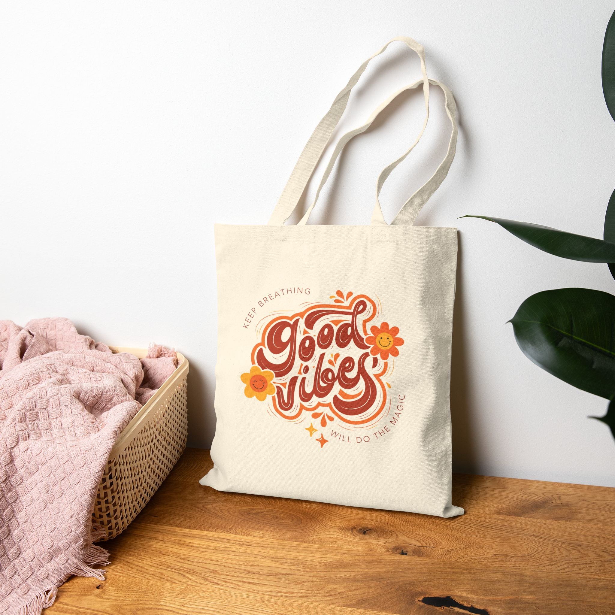 Beige tote bag with 'Good Vibes' design on a wooden surface with a white wall and plant in the background