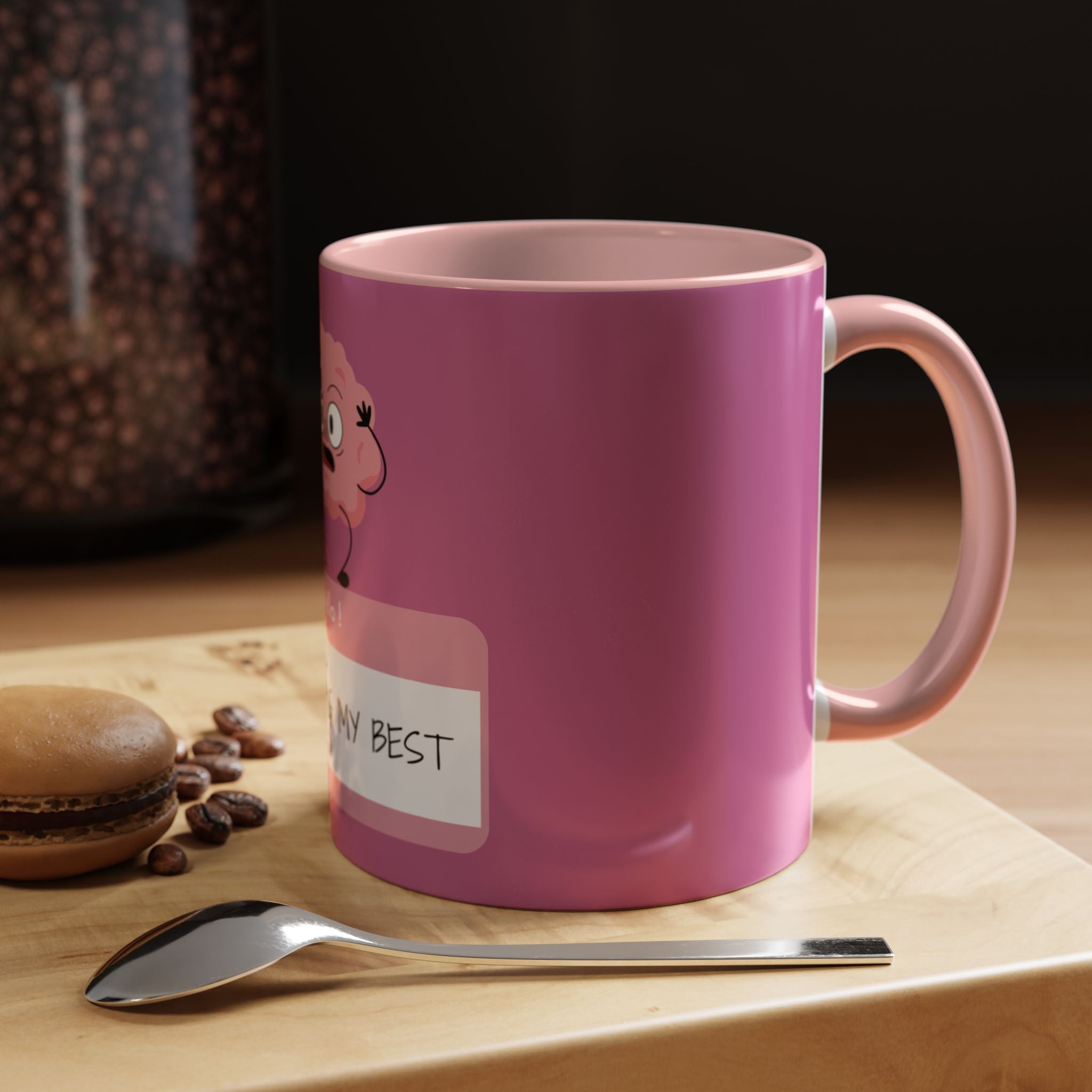 Pink mug with cartoon character and text on a wooden surface with coffee beans and a spoon.