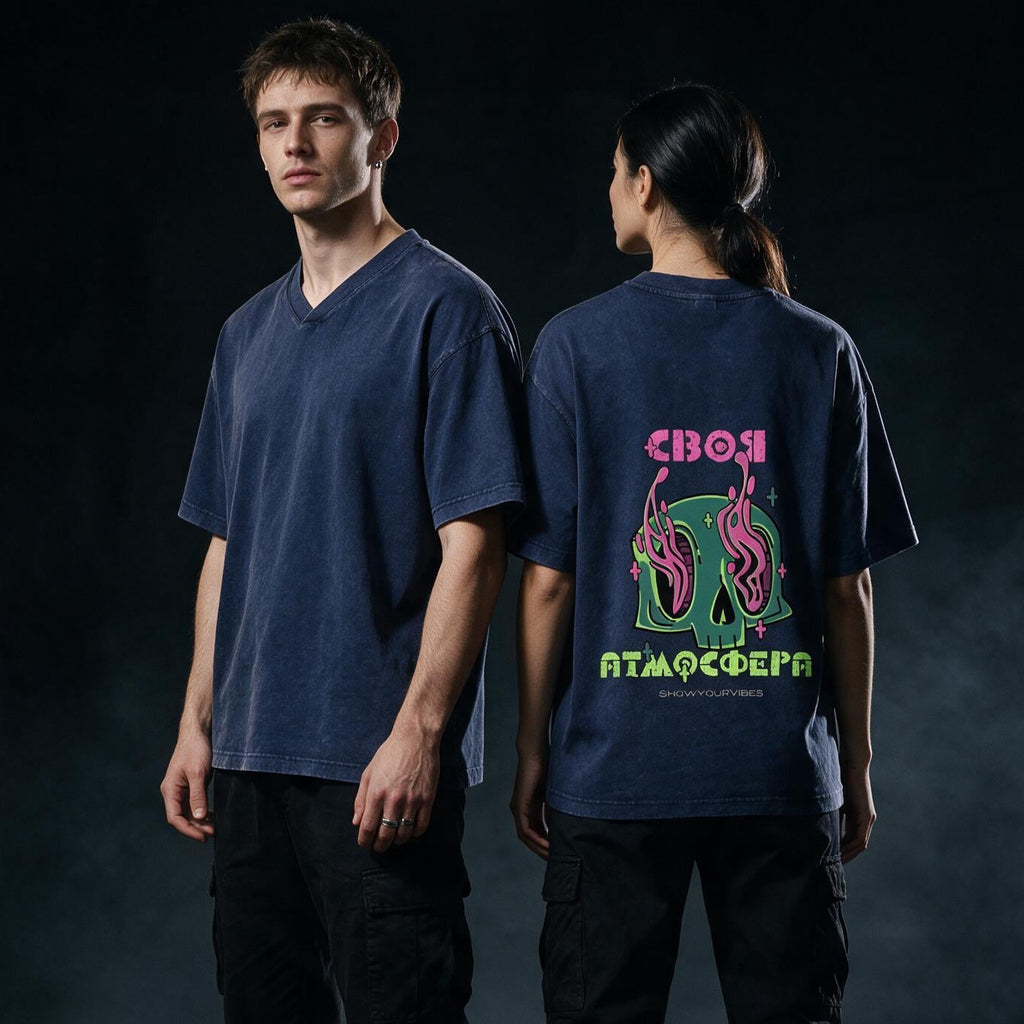 Two models standing in a dark industrial setting, showing a navy blue oversized V-neck t-shirt from the front and back. The back features a colorful graphic with a green skull, pink snakes, and neon text.