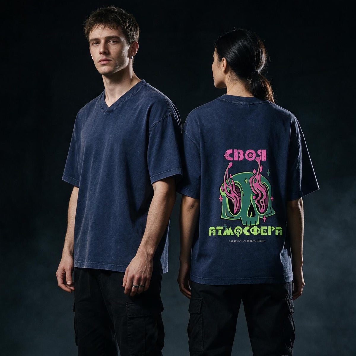 Two models standing in a dark industrial setting, showing a navy blue oversized V-neck t-shirt from the front and back. The back features a colorful graphic with a green skull, pink snakes, and neon text.