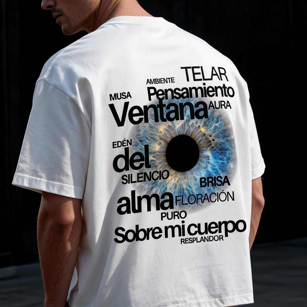 A man wearing a white t-shirt, viewed from the back. The shirt features a large graphic print with a blue and black iris in the center and various Spanish words in black text surrounding it.