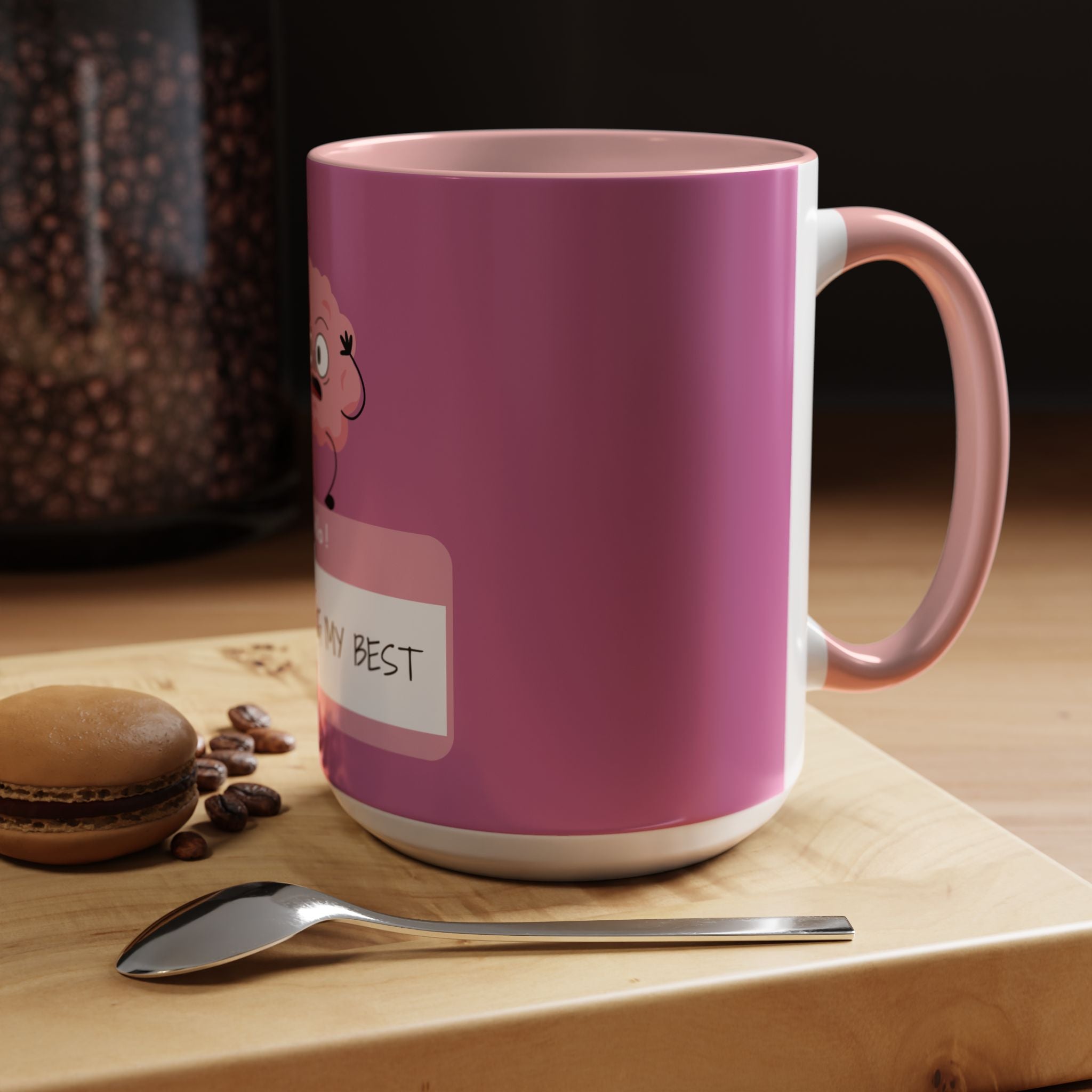 Pink mug with cartoon character and text on a wooden surface with coffee beans and a macaron.