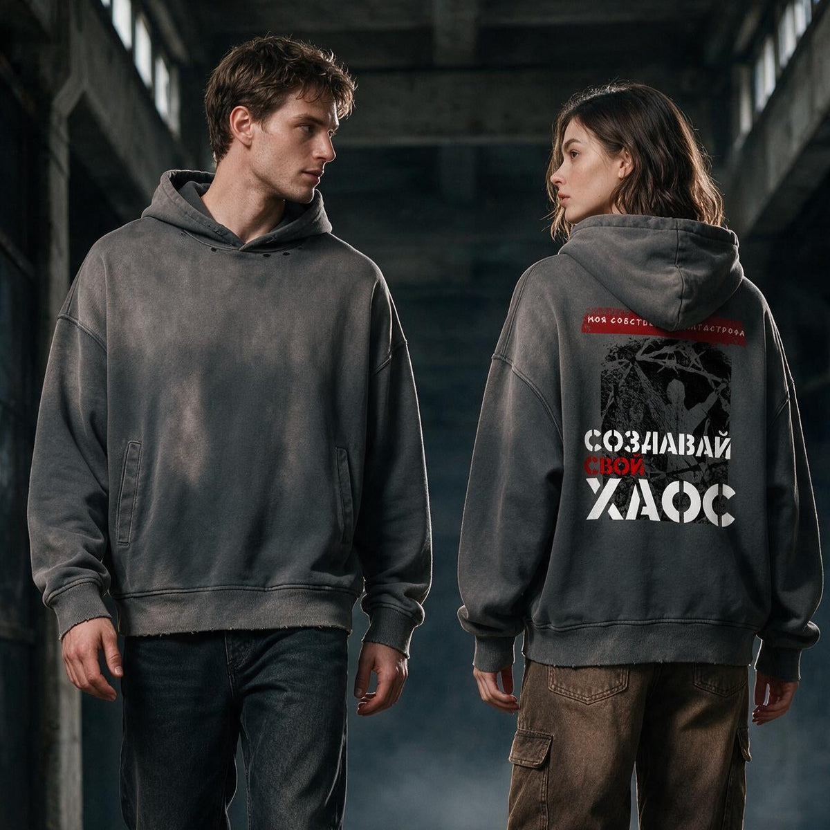Two models walking down a dark, industrial corridor wearing matching dark gray oversized hoodies. The model on the right shows the back of the hoodie, which features a graphic print with a skull and the Russian text 'Удениетик задомой' in white and red.