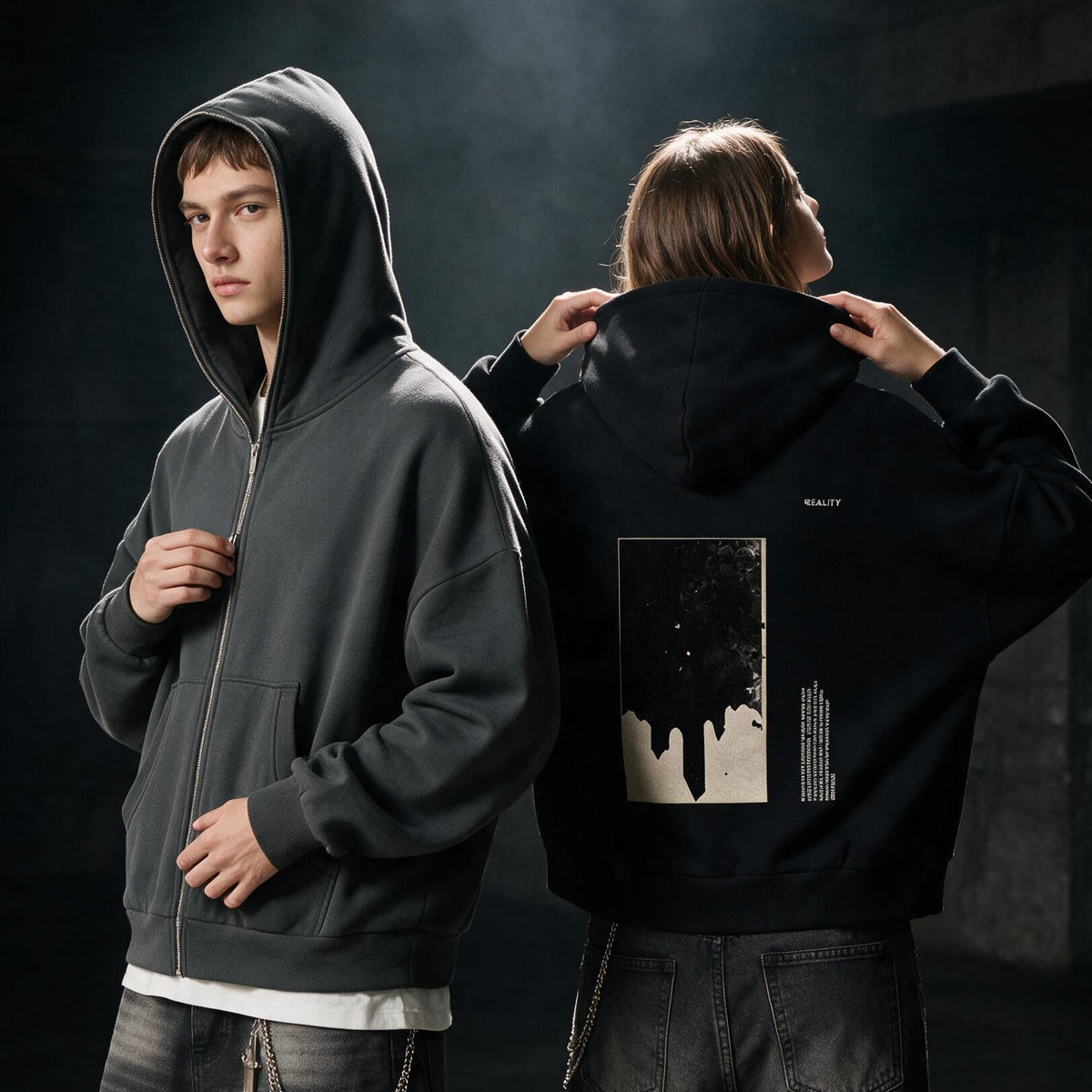 Two models in a dark industrial setting wearing dark hoodies, showing both the front and back views. The front-facing model wears a dark gray zip-up hoodie, while the back-facing model wears a black hoodie with a large graphic print on the back.