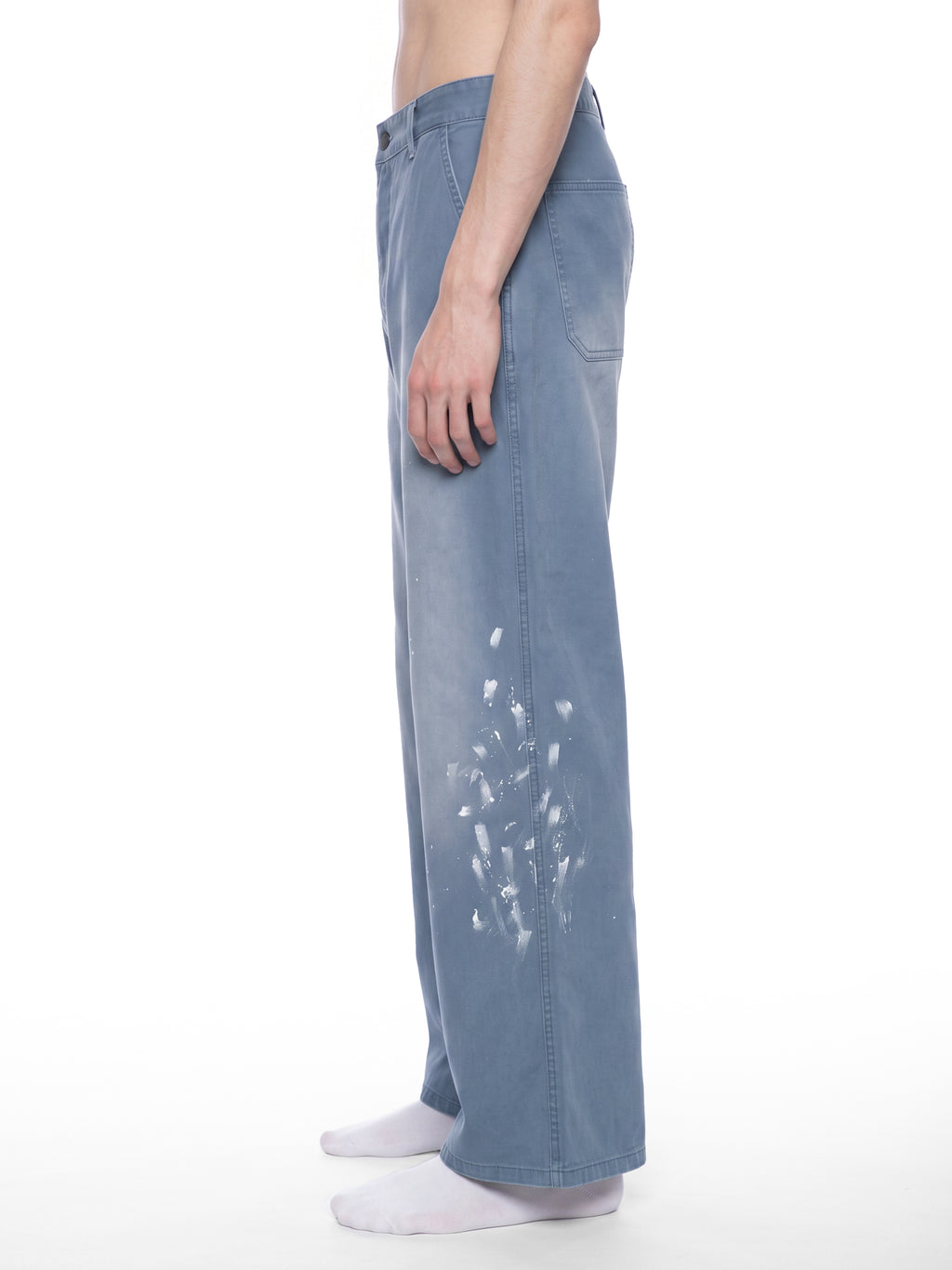 A side-profile view of a model wearing wide-leg, light blue denim pants with white paint splatter details on the lower leg, paired with white socks against a plain white background.