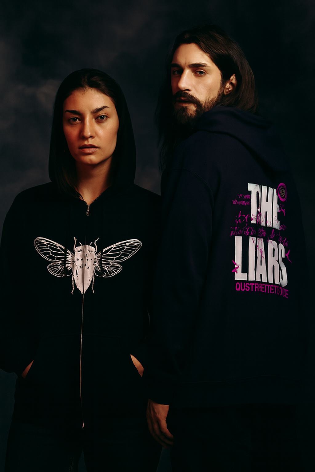 Two people wearing black hoodies with graphic designs on a dark background