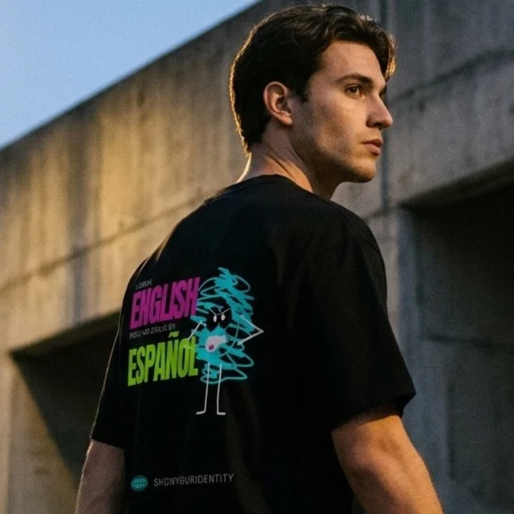 Un hombre con una camiseta negra que lleva un diseño gráfico colorido en la espalda, de pie en el exterior junto a un muro de hormigón al atardecer. El diseño incluye el texto «COPENHAGEN ENGLISH AND SPANISH» en rosa y verde, junto a un personaje de dibujos animados con forma de árbol azul.