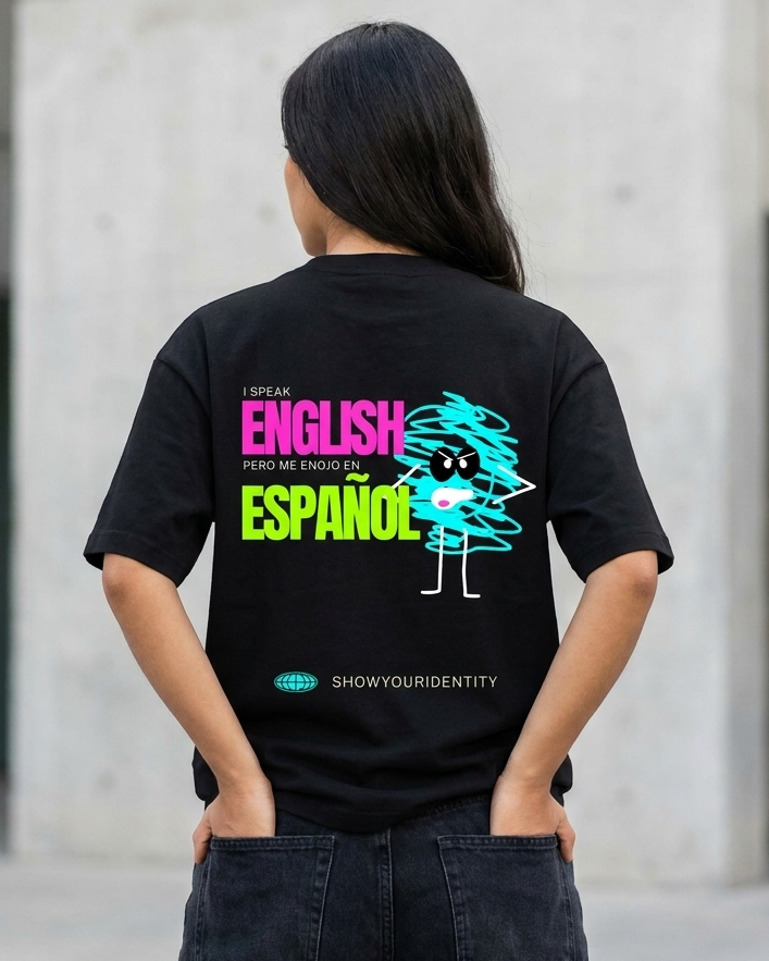 Una persona de pie en el exterior, frente a un edificio de hormigón y vista desde atrás, que lleva una camiseta negra con un diseño gráfico —que incluye un personaje de dibujos animados y el texto colorido «ENGLISH AND ESPAÑOL»— y va vestida con vaqueros oscuros.