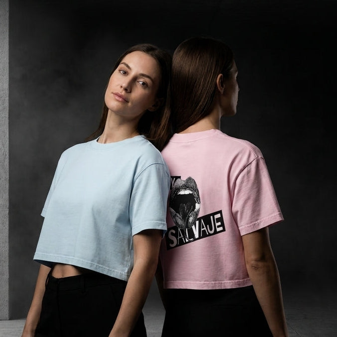 Dos mujeres de pie, espalda contra espalda, en un pasillo oscuro de hormigón; ambas llevan camisetas cortas con estampados gráficos. La mujer de la izquierda viste una camiseta azul claro con un diseño gráfico en negro, y la de la derecha lleva una camiseta rosa con un diseño gráfico en negro y la palabra «SAVAJE».
Camiseta con el diseño gráfico «Stay Salvaje», que presenta una llamativa ilustración de labios en blanco y negro y un mensaje en *spanglish* que celebra el orgullo latino, la confianza, la autoe