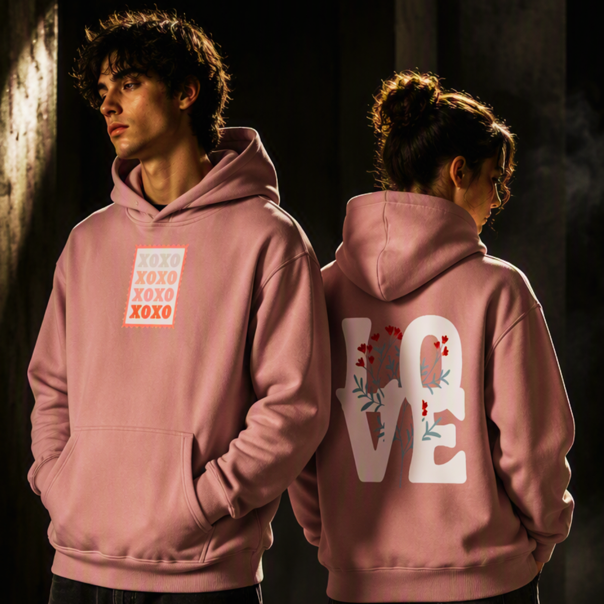 A man and a woman modeling dusty pink hoodies in a dimly lit setting. The man shows the front of the hoodie, which features a square graphic with stacked 'XOXO' text in red and orange.Express your feelings with the ShowYourLove™ XOXO & Floral LOVE Hoodie. 