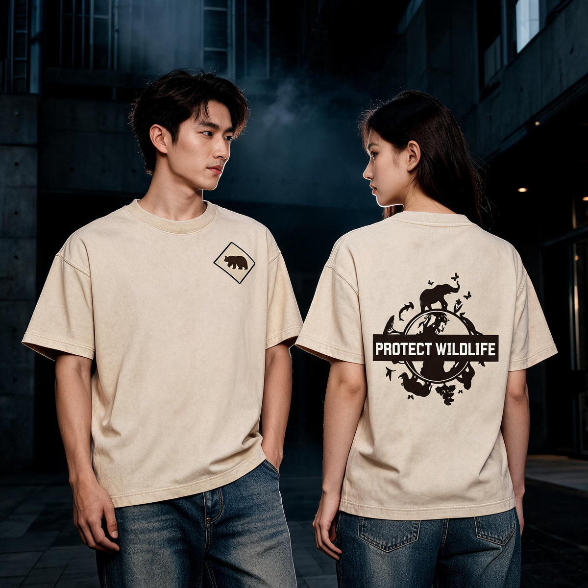 A man and a woman wearing matching beige graphic T-shirts in a dark urban alley. The man wears a small bear logo on the left chest, and the woman wears a large black 'PROTECT WILDLIFE' graphic on the back.