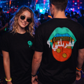 Two people standing together in a club-like environment wearing matching black t-shirts. The woman wears a small green alien graphic on the chest, and the man shows a large back graphic featuring a red tongue with yellow smiley faces, a turquoise mouth, and white Arabic text.